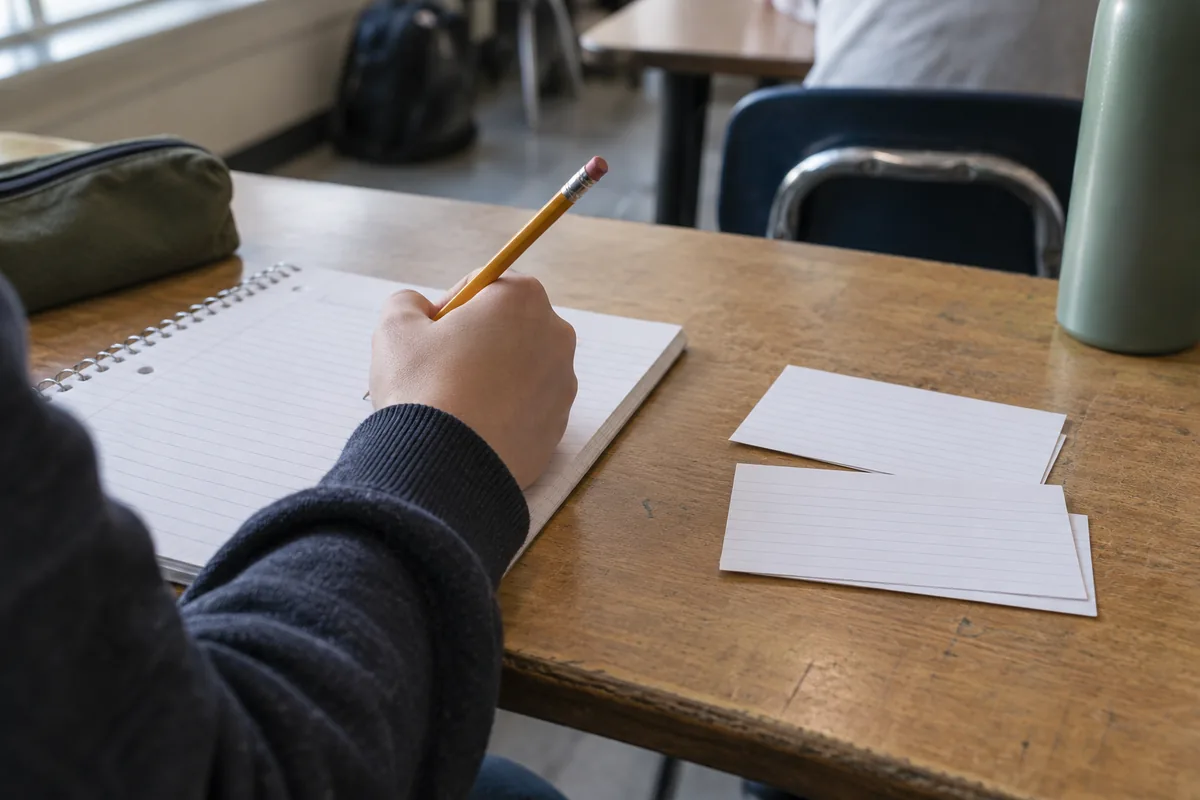 A student’s hand writing in a notebook next to a few blank index cards on a classroom desk.