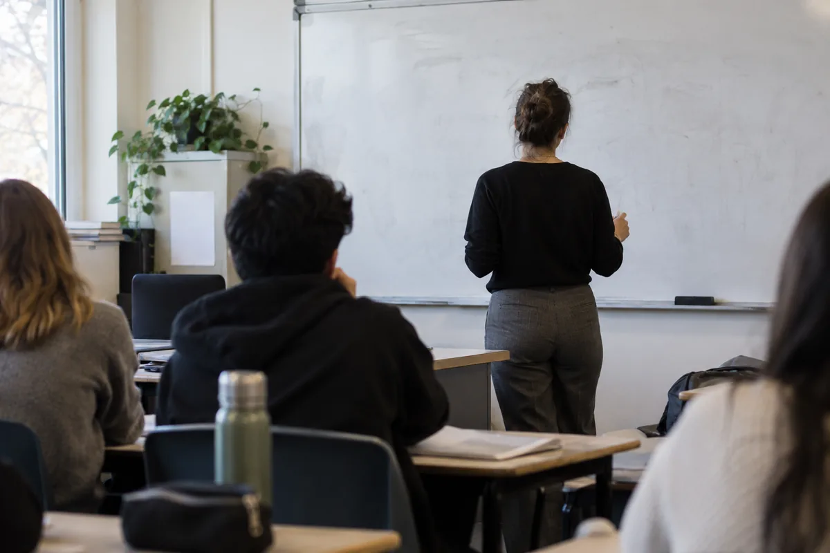 A teacher seen from behind standing near a whiteboard with erased marks in a classroom.